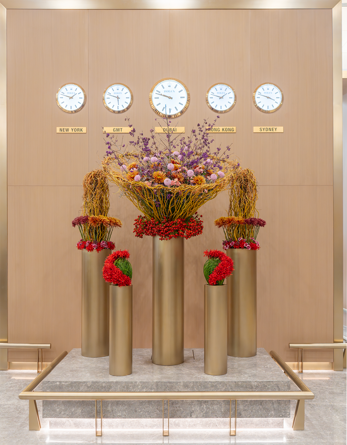 Decorative floral arrangements in tall gold vases against a beige wall with clock faces.