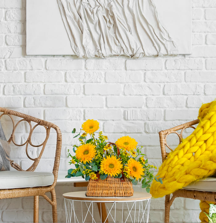 Yellow sunflowers in a woven basket on a small round table with wicker chairs and a white brick wall background.
